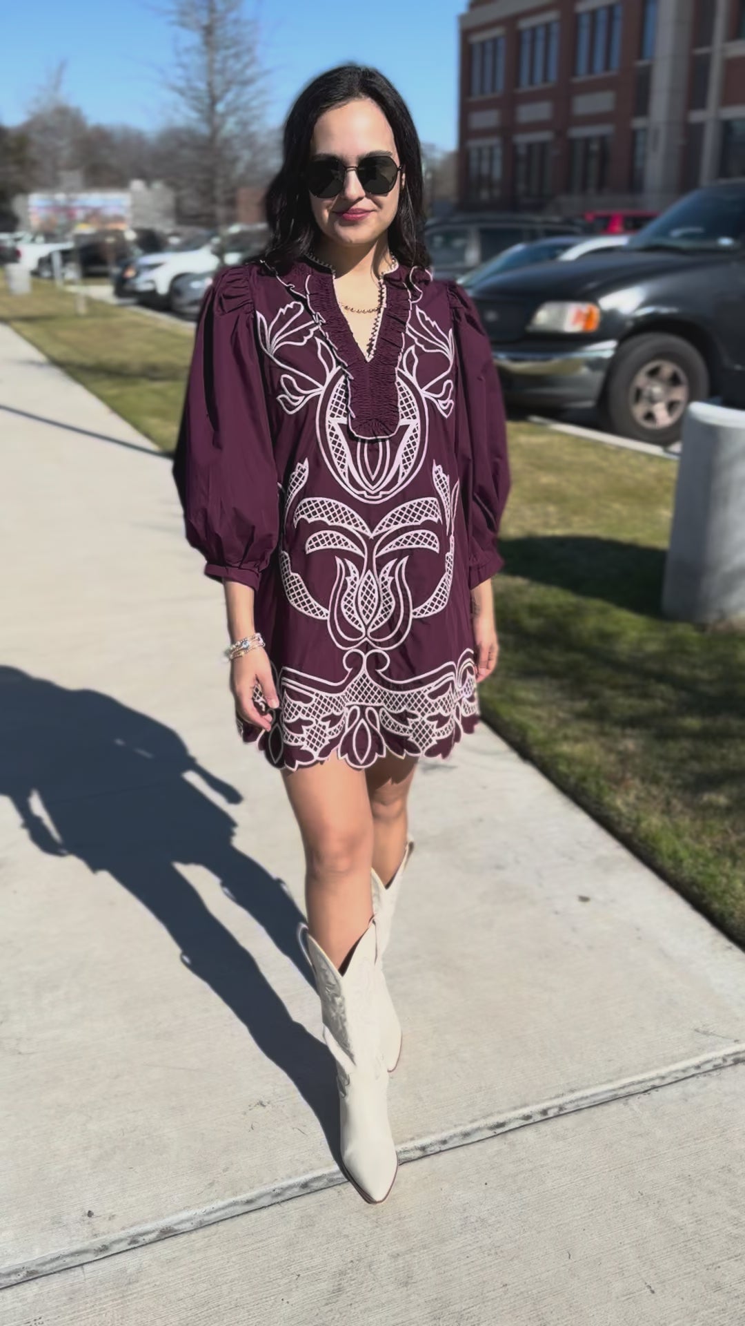 Woman wearing a burgundy embroidered dress with white boots standing outside.