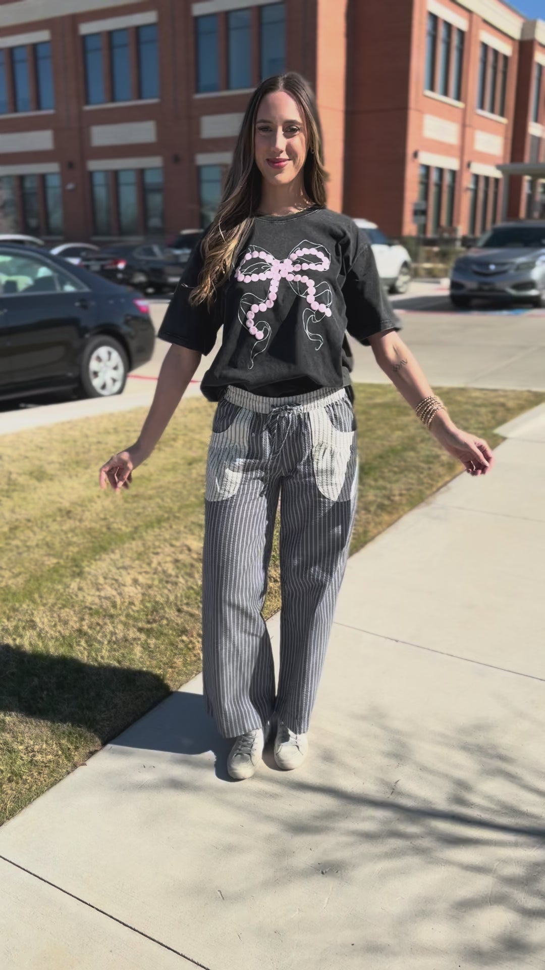 Woman wearing a black t-shirt with a pink bow design and striped pants outside.