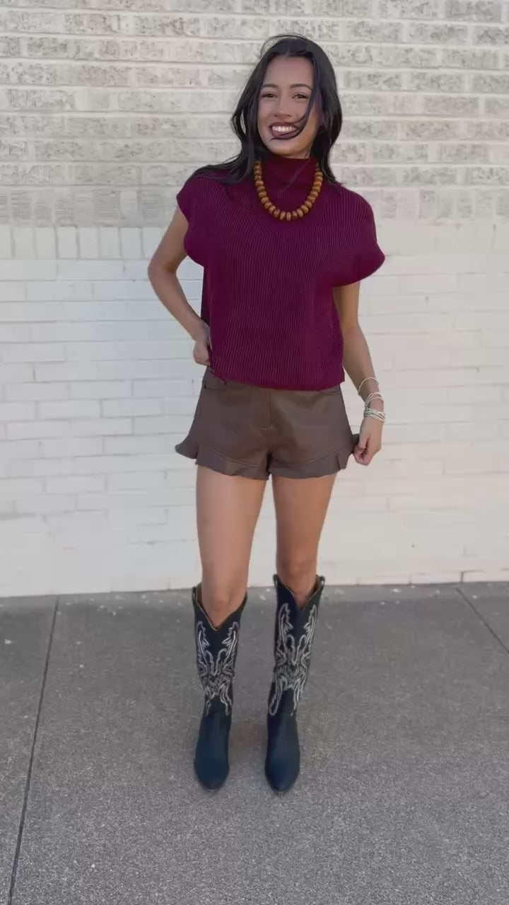 A woman posing outdoors on a sidewalk next to a white brick wall, wearing a burgundy knit t-shirt, brown leather shorts, and black western boots with white embroidery.