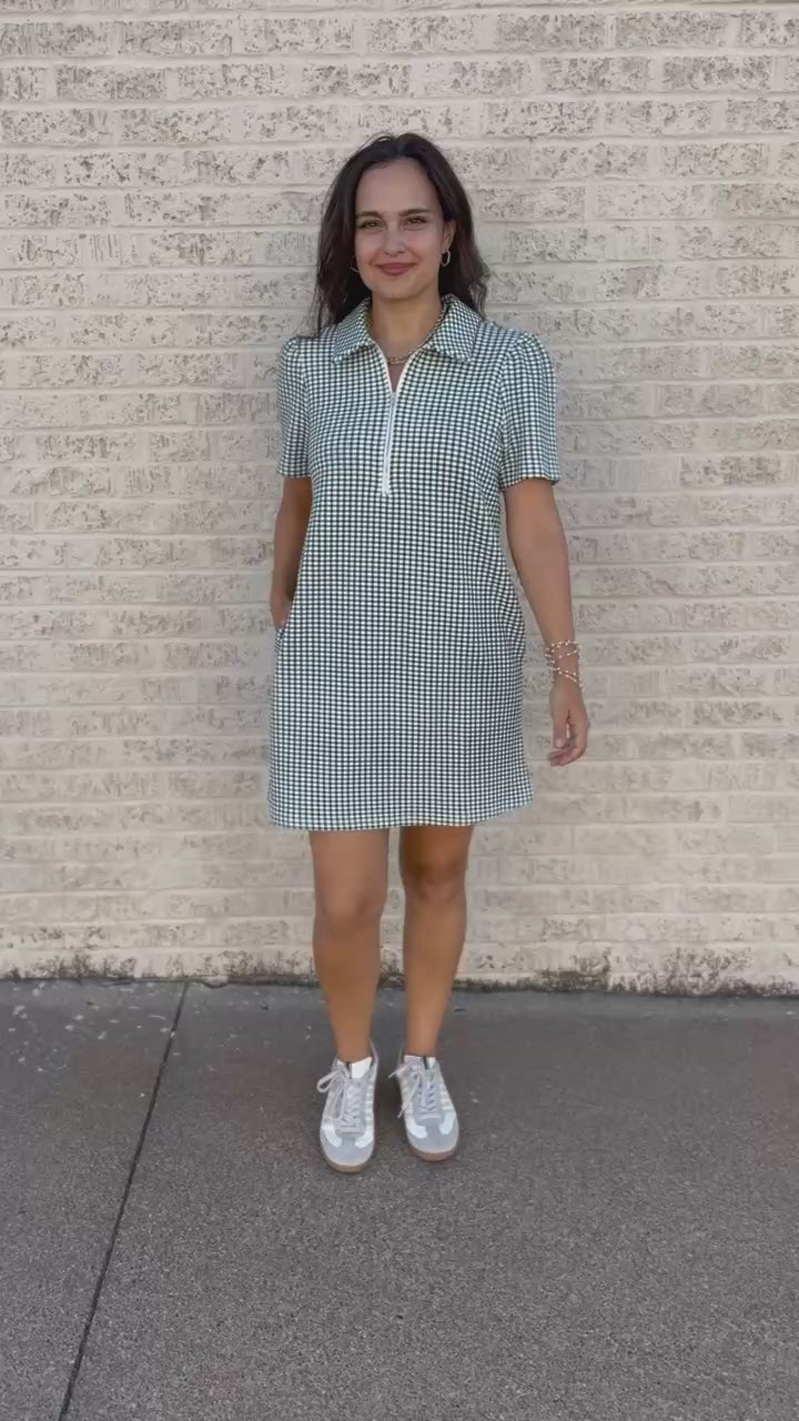 A woman wearing a short-sleeve gingham mini dress with a half-zip collar and side pockets, walking outdoors on a paved path