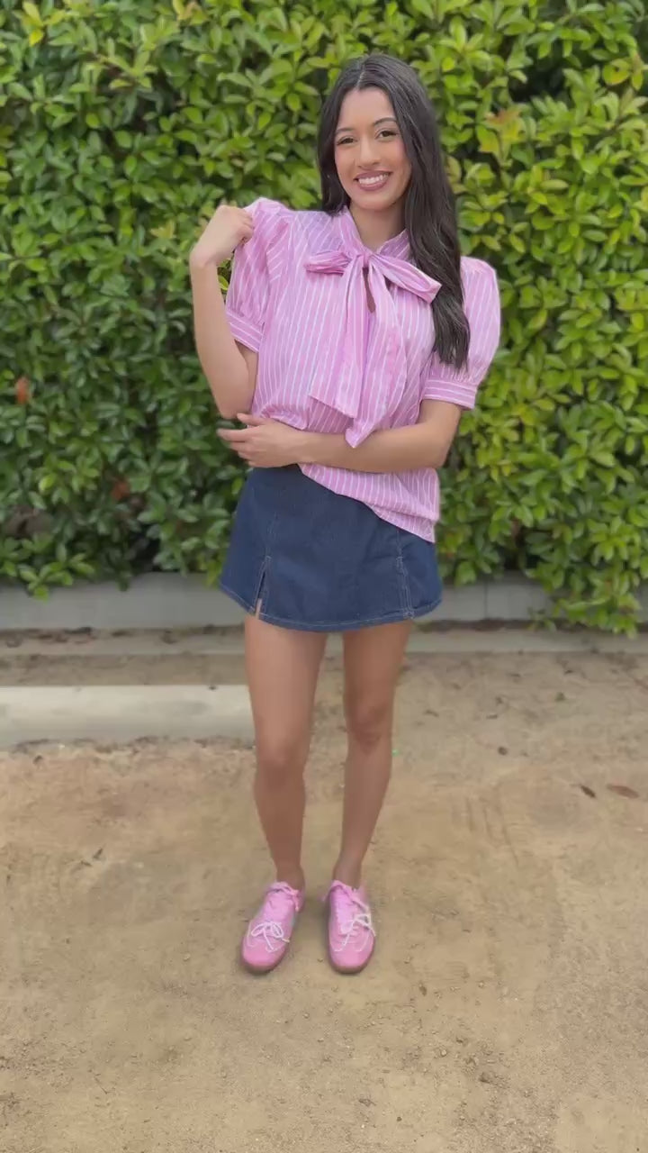 Woman wearing a pink striped shirt, denim skirt, and pink shoes standing outdoors.