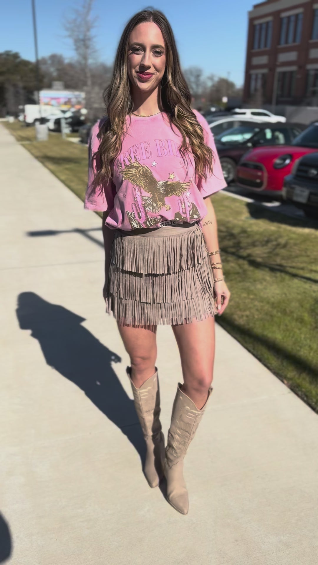 Woman wearing a pink blouse with a graphic design and a beige fringe skirt, standing on a sidewalk.