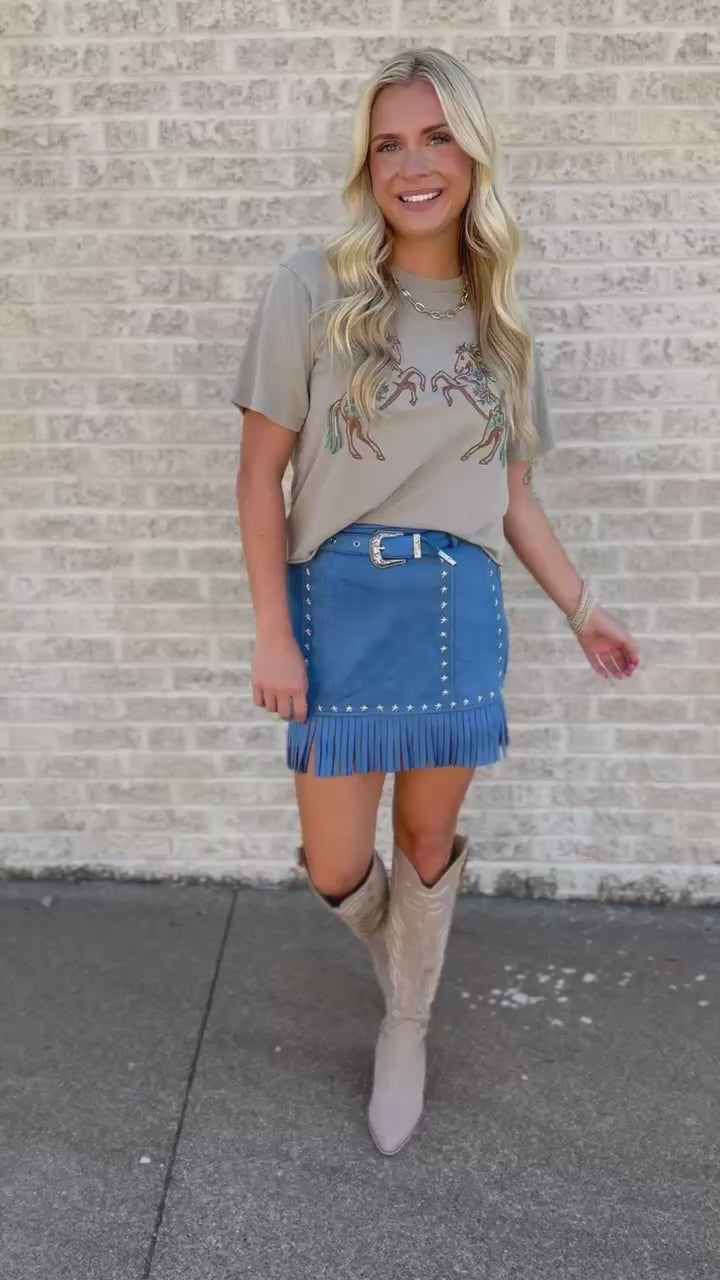 A woman stands outdoors against a light brick wall wearing a beige graphic T-shirt, a blue denim mini skirt with fringe and silver studs, and tall beige cowboy boots.