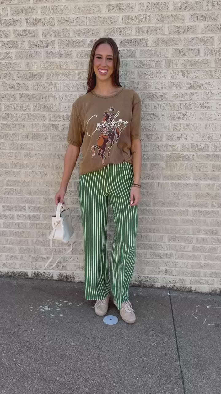 A woman walking outdoors on a sidewalk next to a light brick wall, wearing a brown graphic t-shirt and green and white vertically striped wide-leg pants, carrying a white shoulder bag.