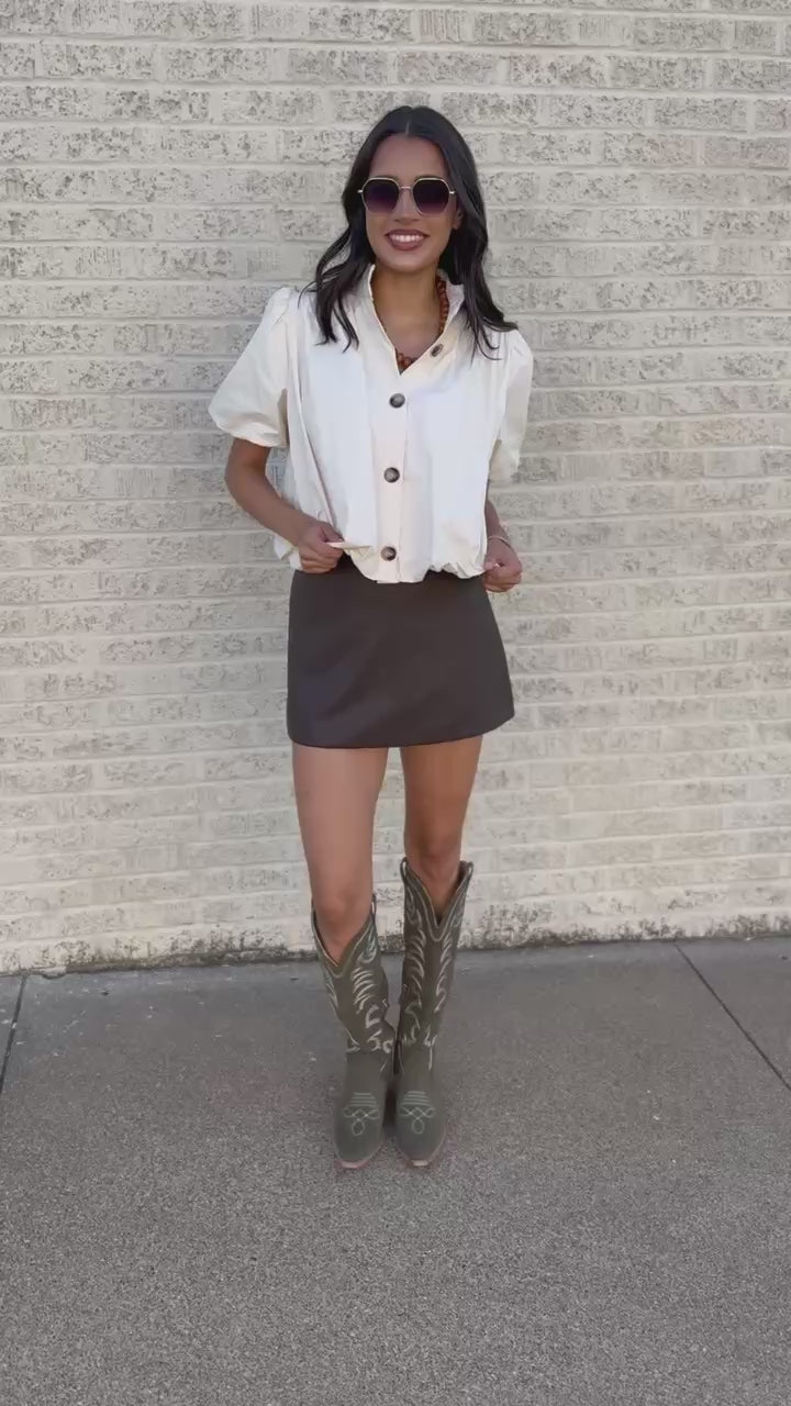 A woman wearing a cream puff-sleeve button-up blouse, a brown mini skirt, and olive green cowboy boots with white stitching, standing outdoors on a paved surface.