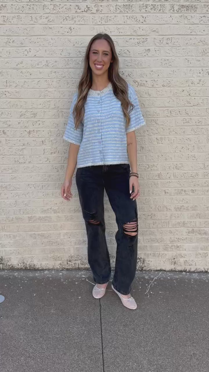 A woman stands outdoors against a white brick wall wearing a light blue and white striped short-sleeve button-up top, black distressed jeans, and white sneakers.