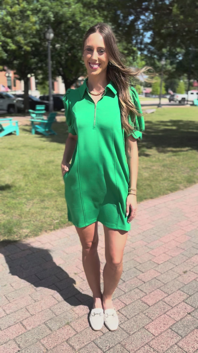 A woman wearing a bright green short-sleeve polo-style mini dress with puff sleeves and a front zipper, walking on a sidewalk 
