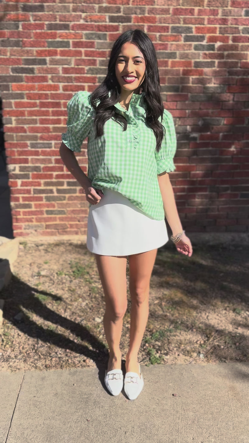 Woman wearing a green checkered blouse, white skirt outside in front of a brick wall.
