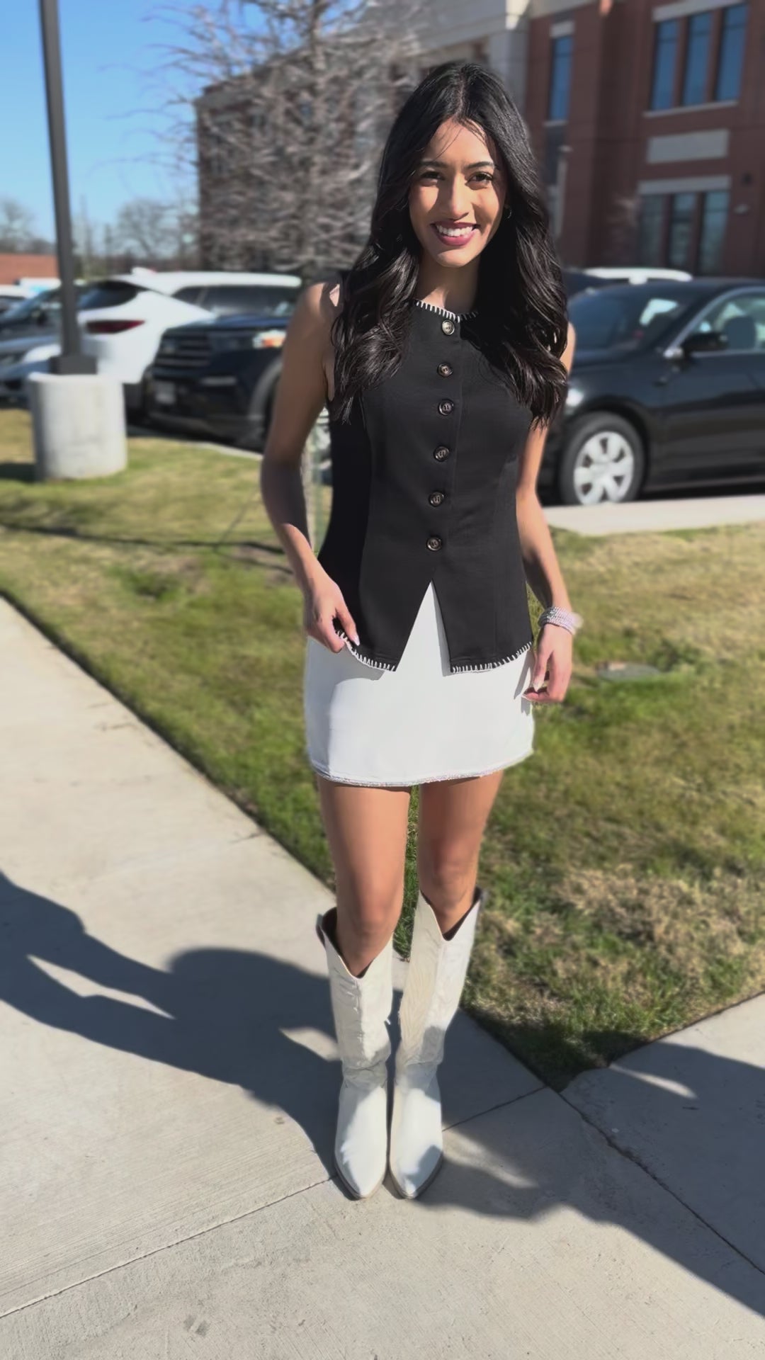 Woman wearing a black vest over a white dress standing outside.