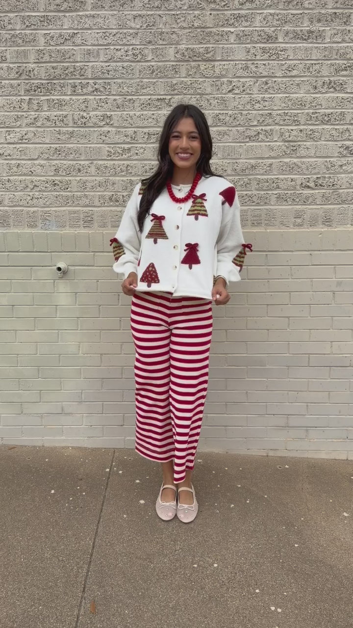 Brunette model wearing the Merry Little Christmas Tree Cardigan in cream featuring red and green Christmas tree patterns, decorative red bows, and front buttons, styled with the Candy Cane Lane Striped knit pants from Southern House Clothing