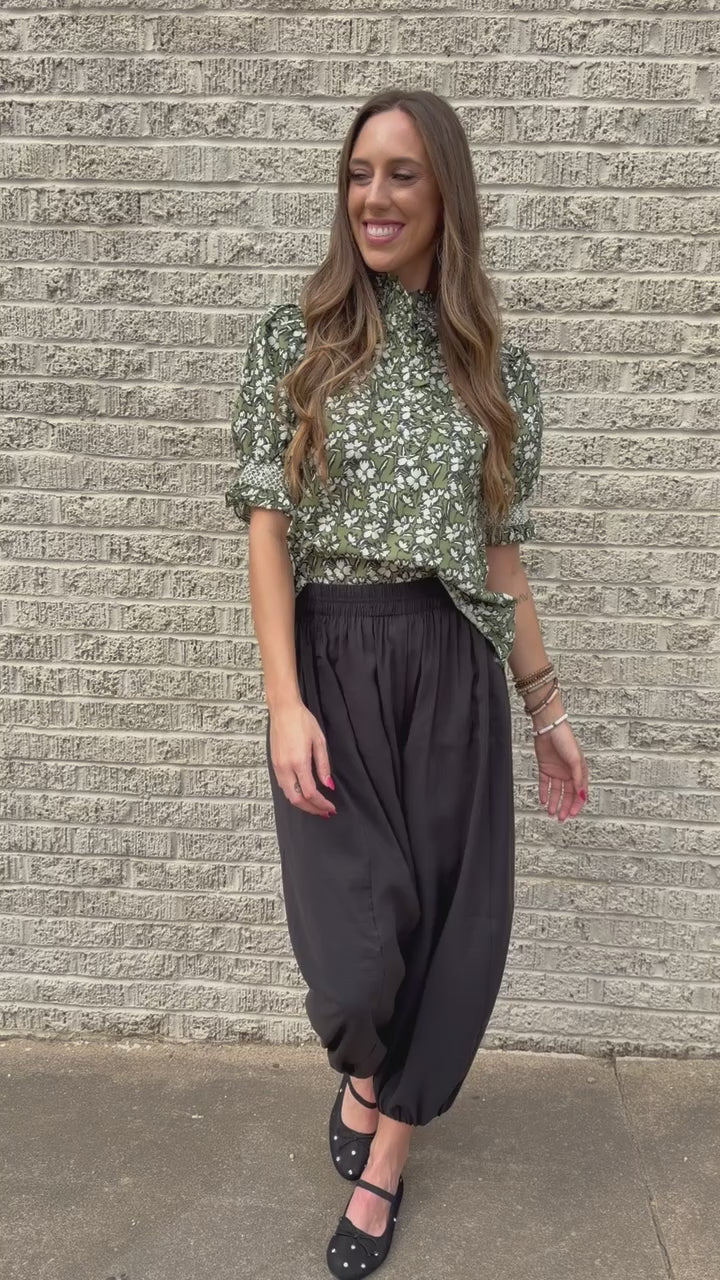 Blond model standing outside wearing a green blouse and black plants with bracelets and black ballet flats.