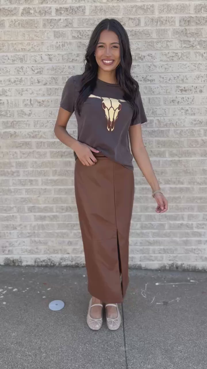 A woman stands on a sidewalk next to a white brick wall, wearing a dark brown graphic t-shirt with a cow print and a brown faux-leather midi skirt.