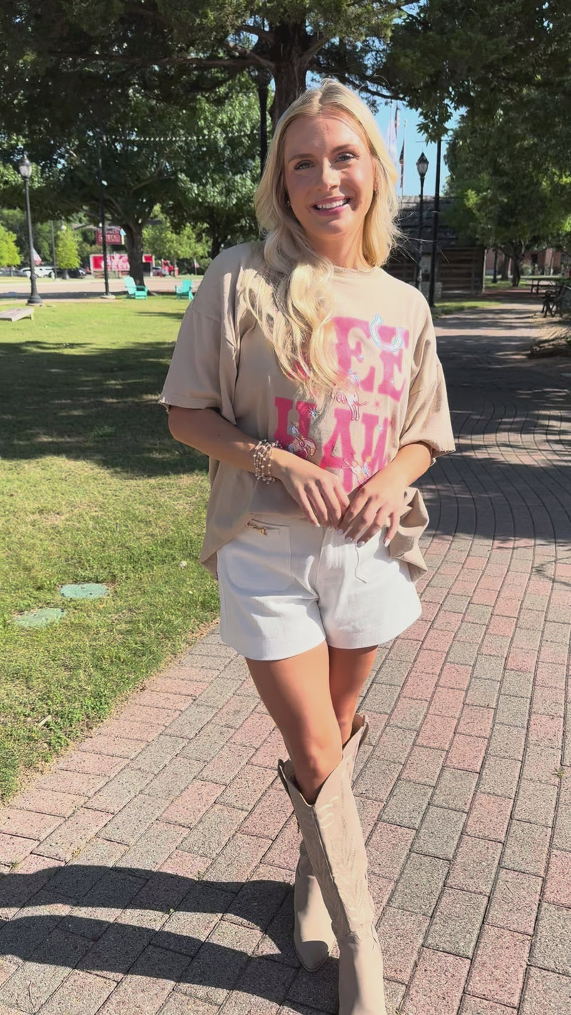 A woman wearing an oversized beige graphic T-shirt with pink 'YEE HAW' lettering and carousel horse illustrations, paired with white shorts.