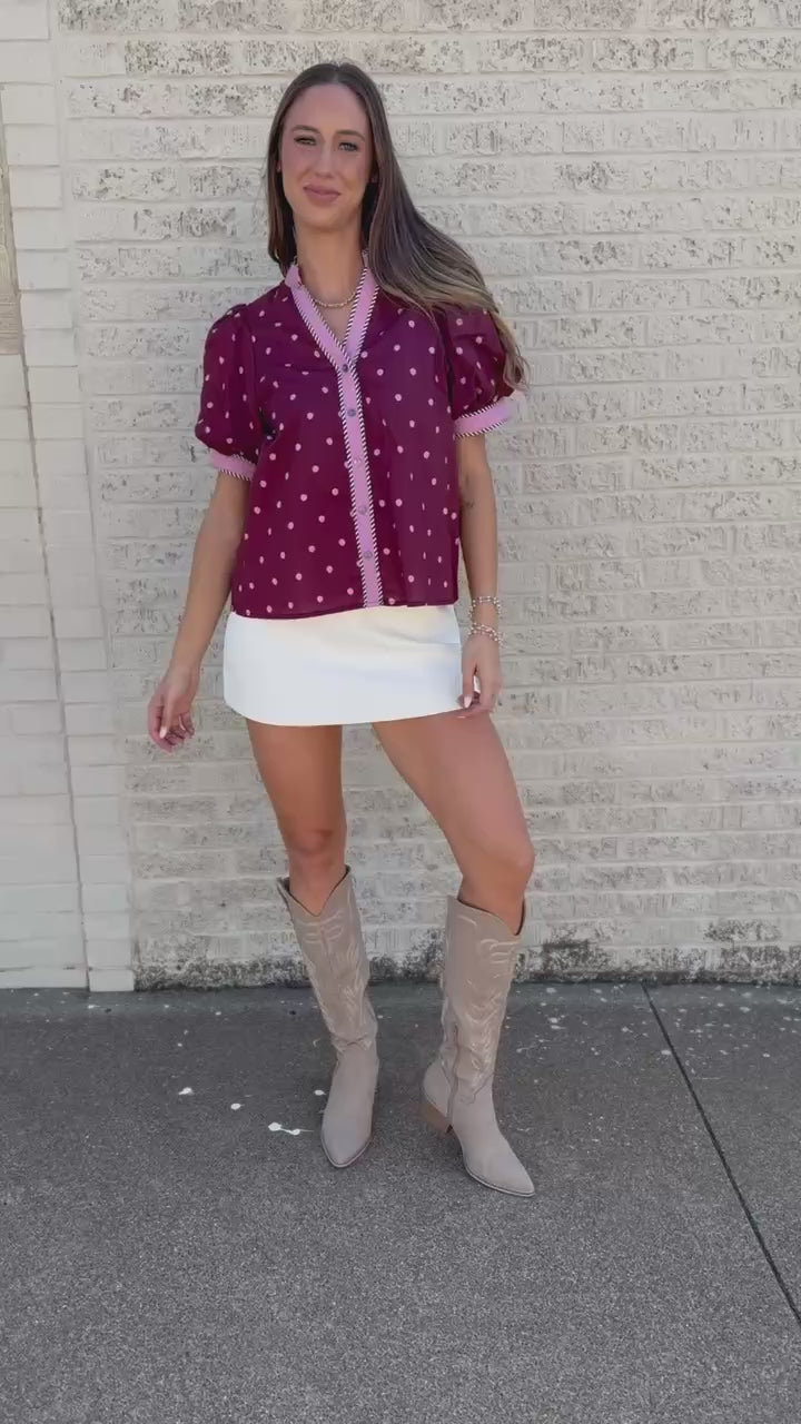 A woman stands on a sidewalk next to a light-colored brick wall, wearing a short-sleeve burgundy button-up blouse with white polka dots and white trim, a white mini skirt, and beige cowboy boots.