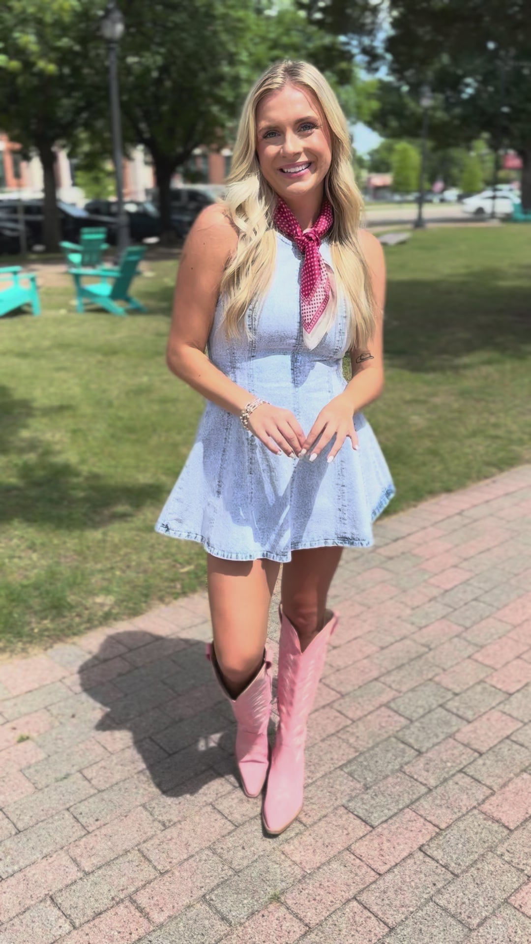 Blonde model wearing a light denim romper with pink cowboy boots, a ruby red scarf, standing outside.