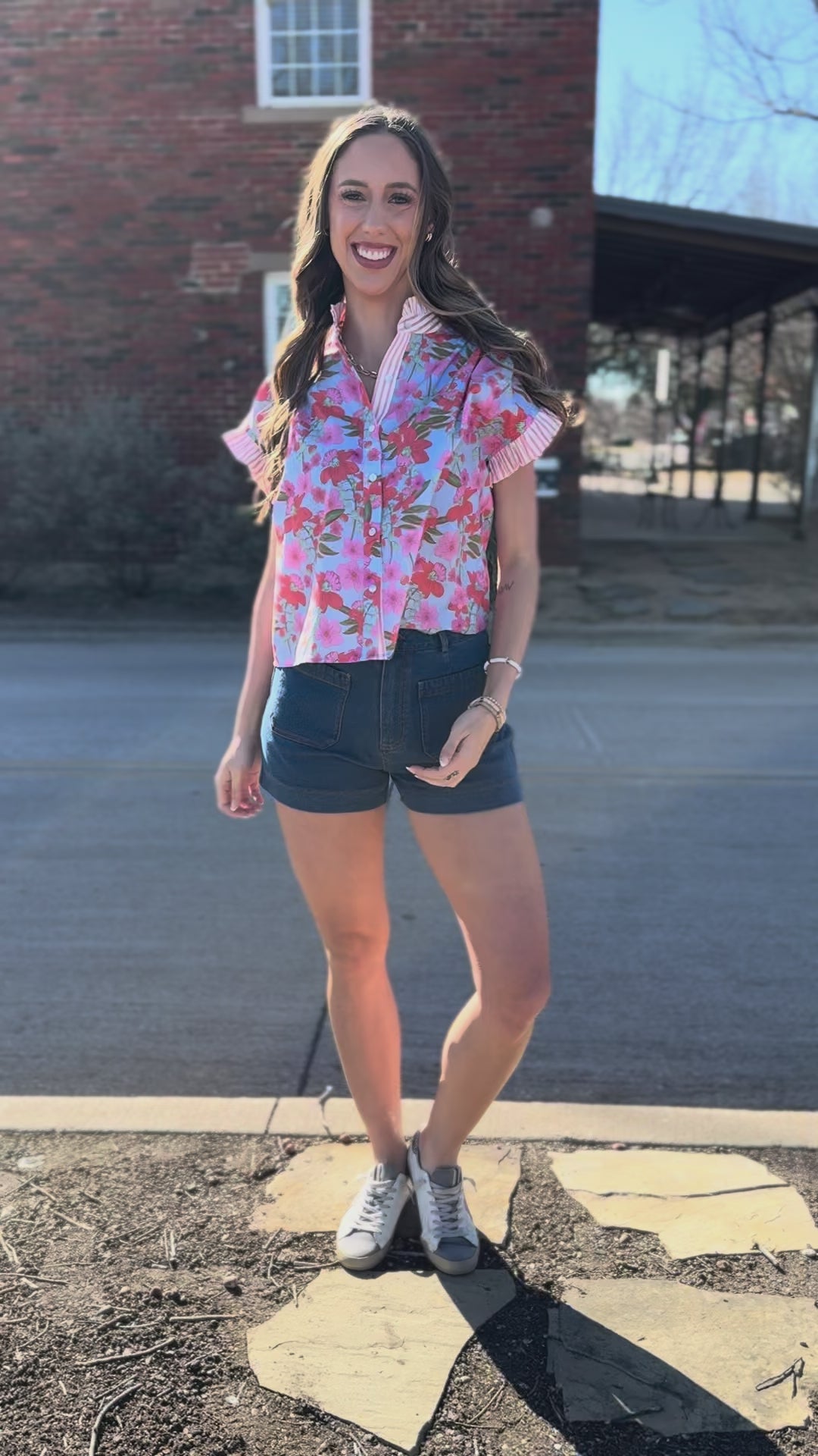 Woman wearing a floral blouse and denim shorts standing on a sidewalk.