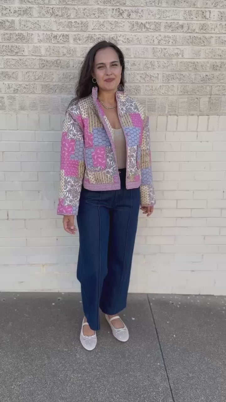 A woman stands outdoors against a light brick wall wearing a pastel patchwork quilted jacket with pink, blue, and beige panels, dark wide-leg jeans, a beige top, and light beige flats.