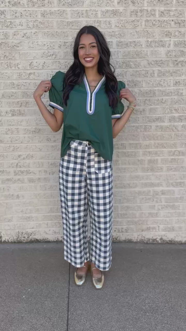 A woman modeling a green short-sleeve blouse with white trim and blue-and-white gingham wide-leg pants, standing against a light brick wall.