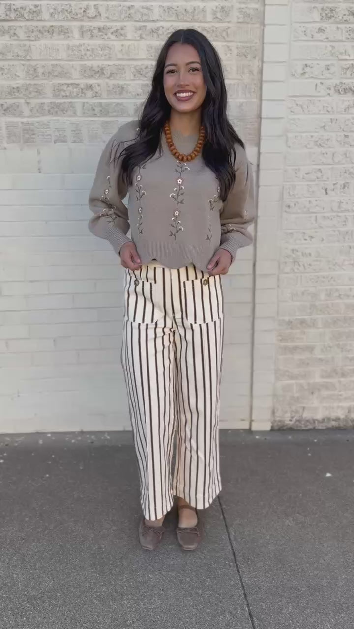 A woman wearing a beige knit sweater with floral embroidery, cream and black striped wide-leg pants, and brown flat shoes, standing on a sidewalk next to a white brick wall.