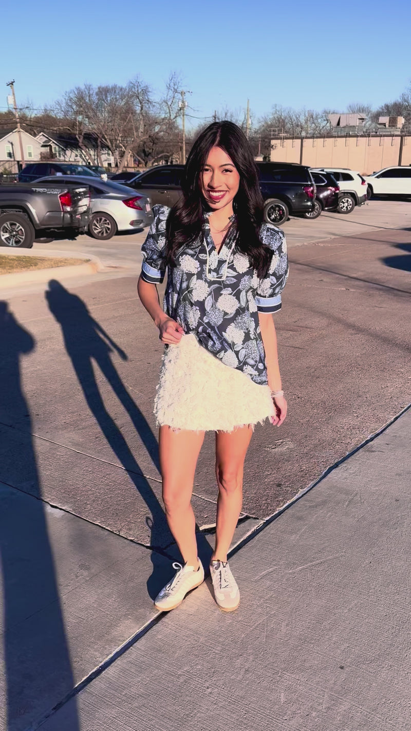 Woman wearing a floral blouse and white skirt standing in a parking lot.