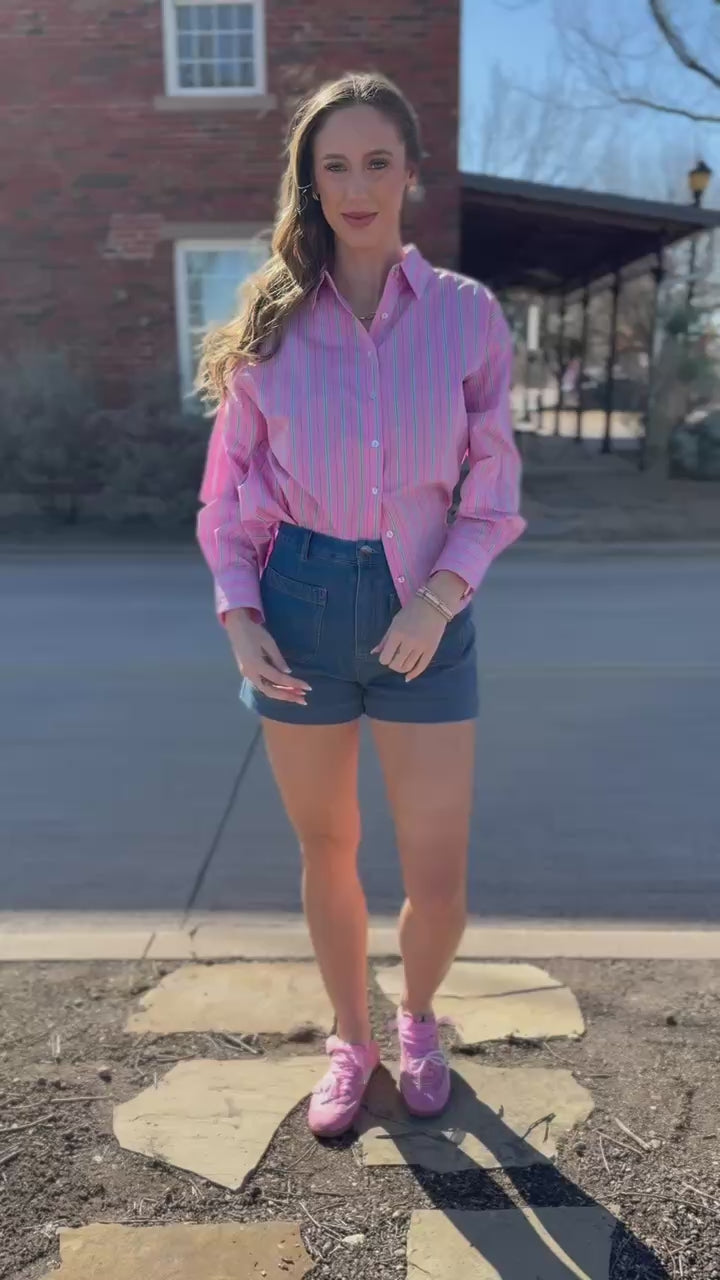 Blonde model wearing a striped pink and green top, denim shorts, and pink shoes outside.