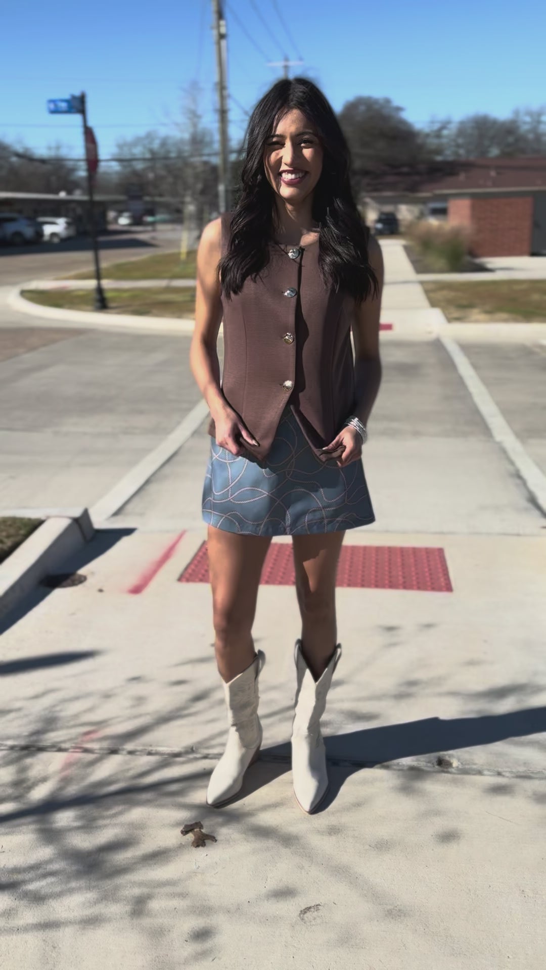 Woman wearing a brown sleeveless top with gold buttons and a blue patterned skirt.