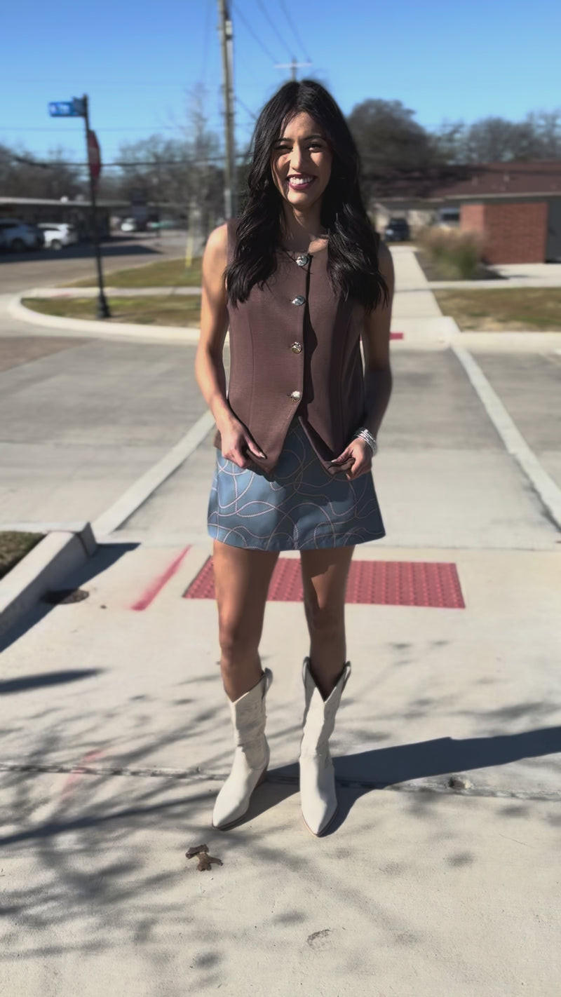 Woman wearing a brown sleeveless top with gold buttons and a blue patterned skirt.