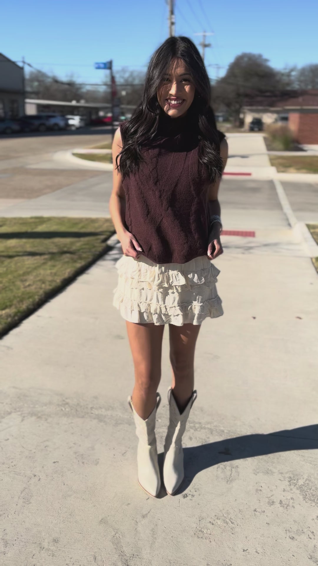 Woman wearing a brown sleeveless sweater and white ruffled skirt outside. 