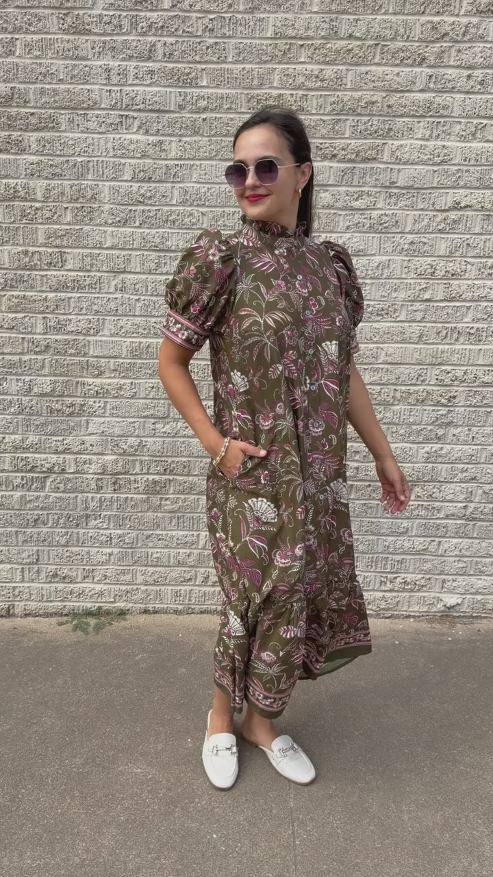 Brunette model wearing an olive dress with patterns standing outside. Southern House Clothing