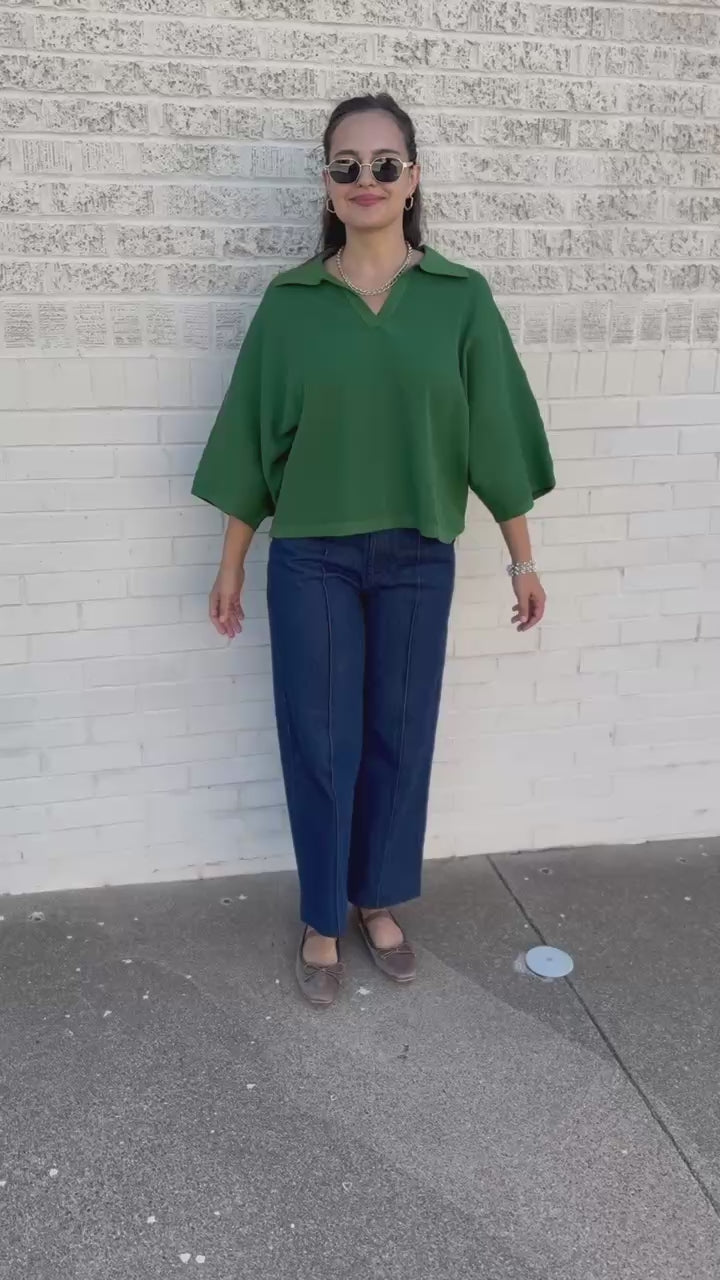 A woman wearing a vibrant green short-sleeve collared top and dark blue jeans, standing outdoors next to a building.