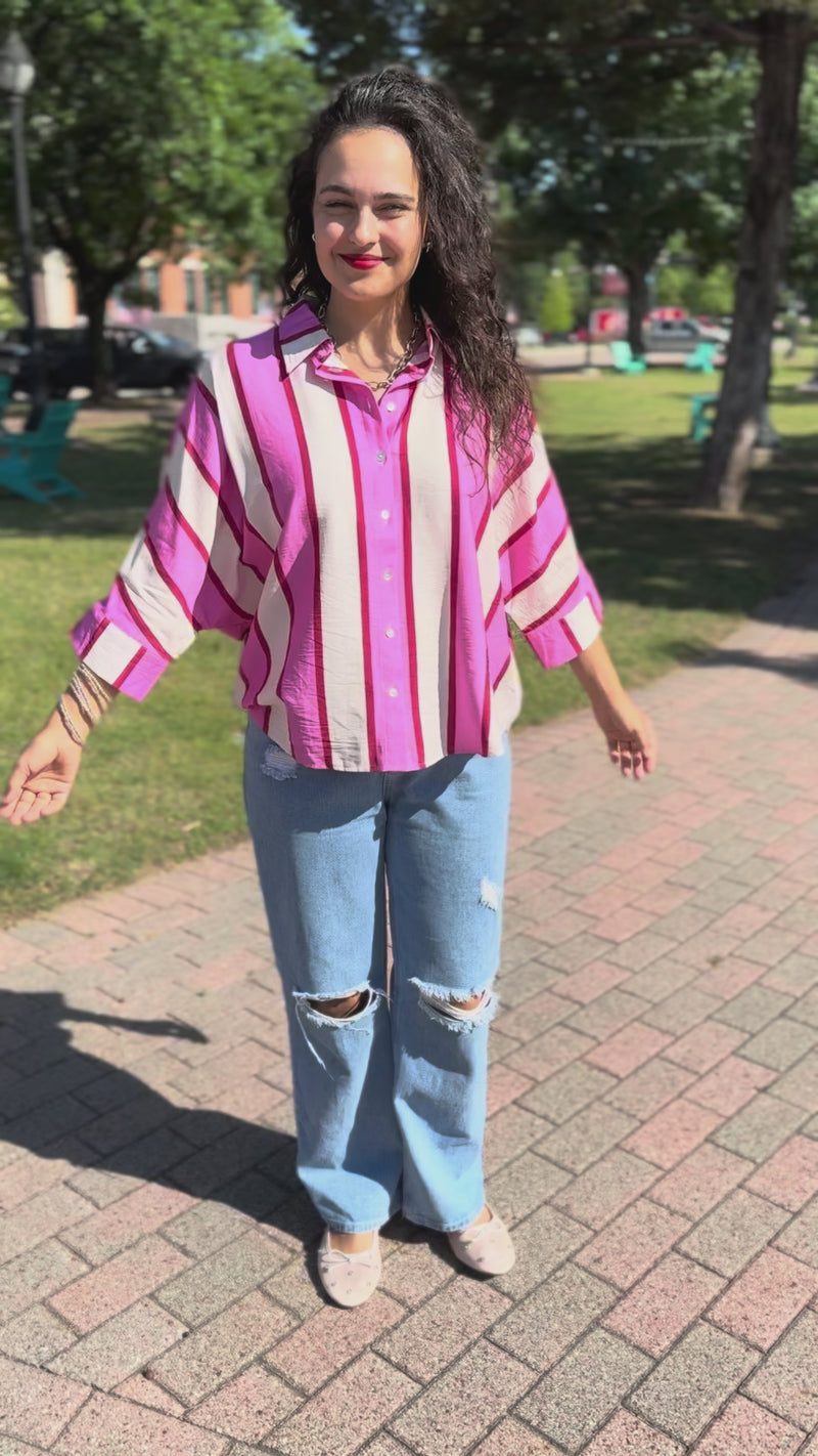 A woman stands outdoors on a sidewalk wearing a pink and white striped long-sleeve blouse, light-wash distressed jeans, white flats.
