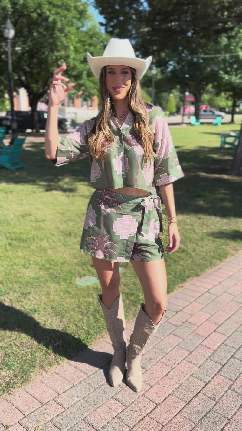 A model wearing a green patterned skirt with palm tree design and matching blouse standing outside.