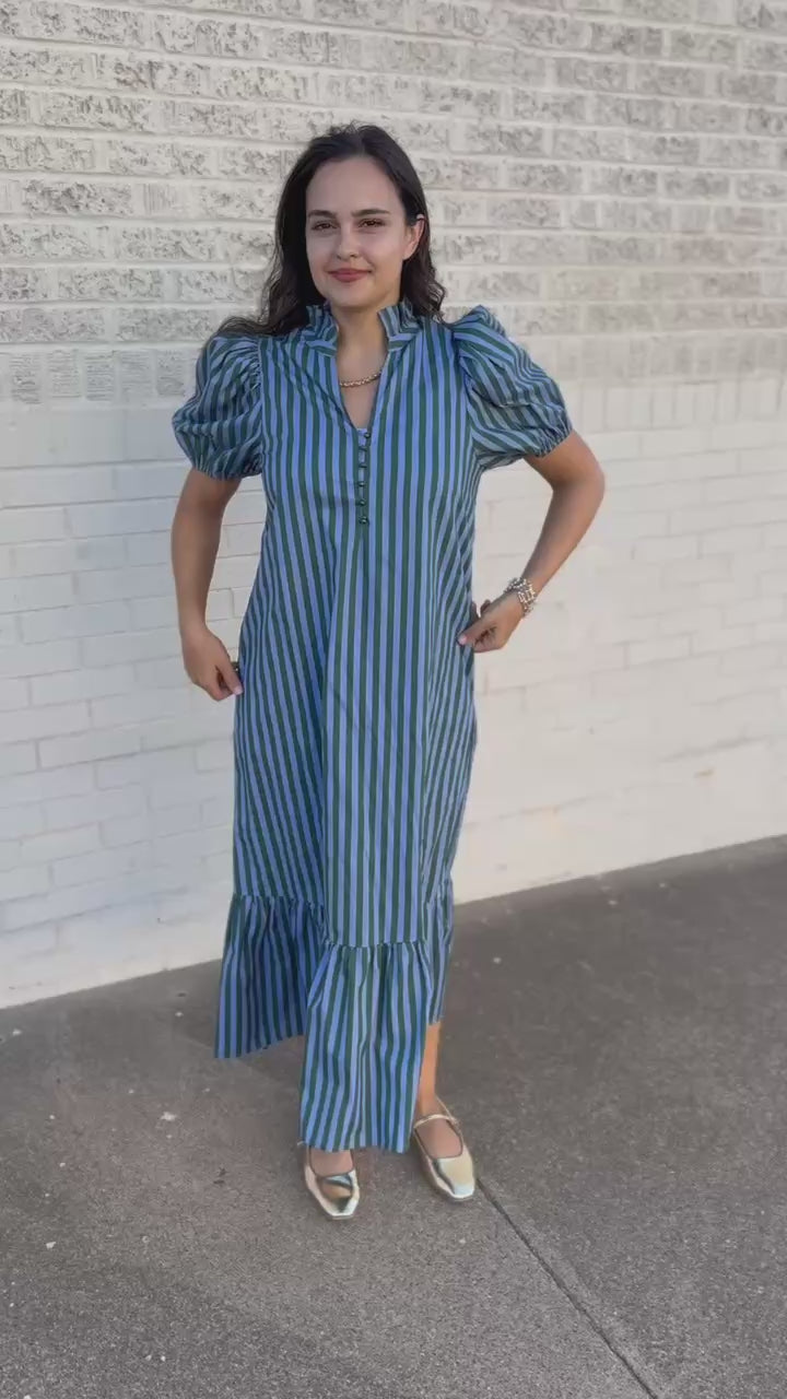 A woman wearing a blue and white vertically striped midi dress with puff sleeves and a ruffled hem, walking outdoors on a sidewalk next to a building.