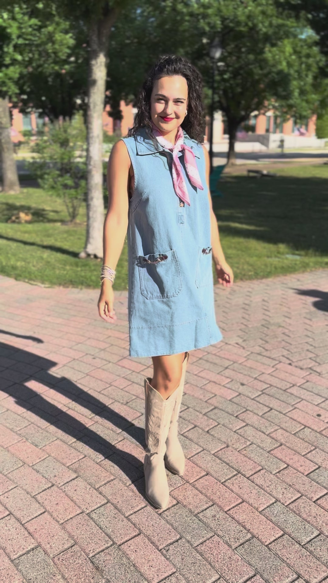 Woman wearing a light blue dress with a pink scarf and beige boots standing outside.