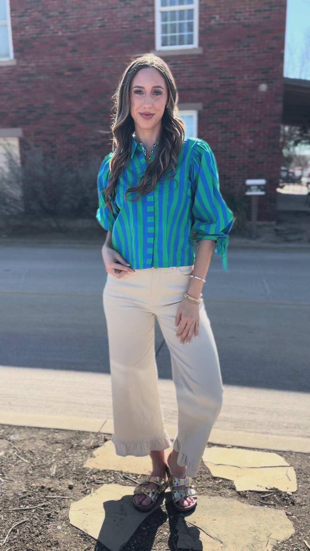 Woman wearing a blue and green striped blouse with tied sleeves, ecru pants standing outside.