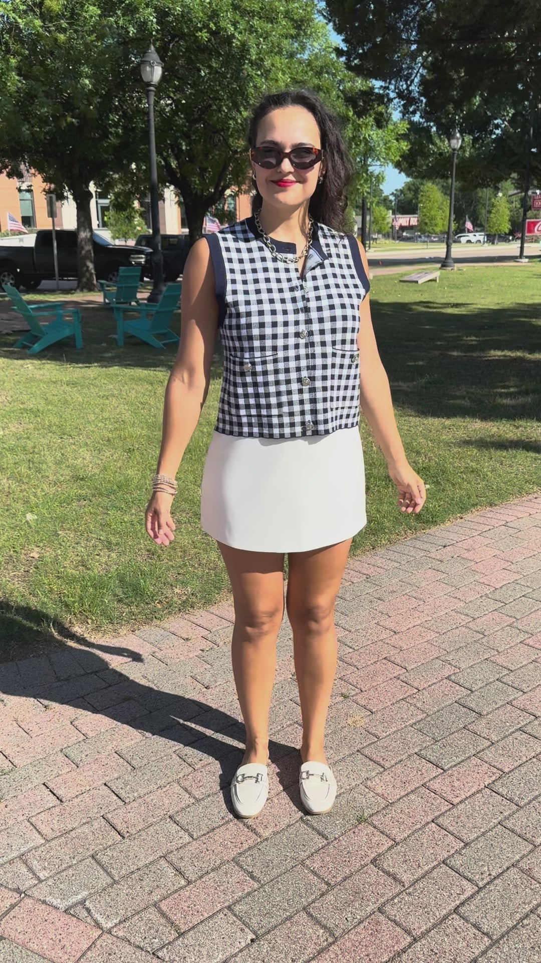 A woman modeling a sleeveless navy and white gingham top with a white mini skirt, white loafers, standing outdoors