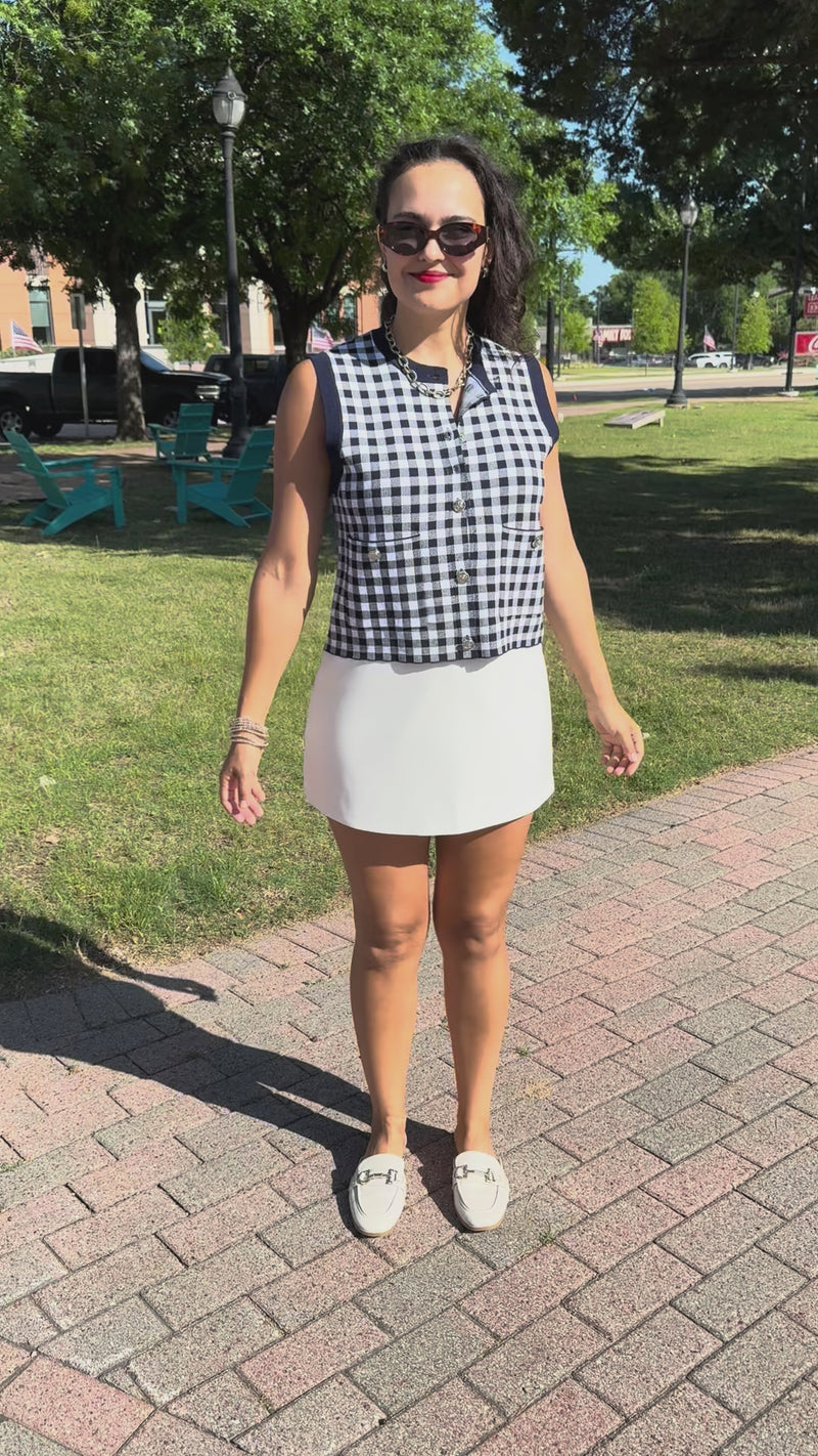 A woman modeling a sleeveless navy and white gingham top with a white mini skirt, white loafers, standing outdoors