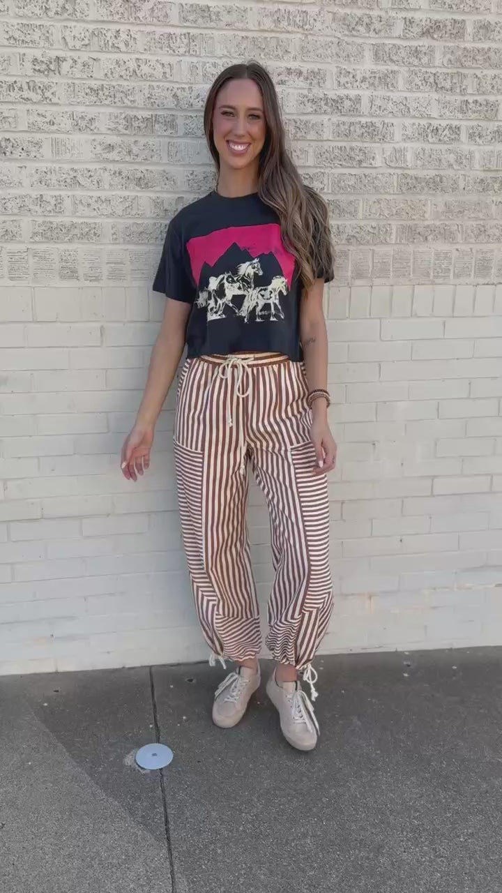 A woman stands outdoors on a sidewalk next to a building, wearing loose brown and white vertically striped drawstring pants, a black graphic t-shirt, and beige sneakers.