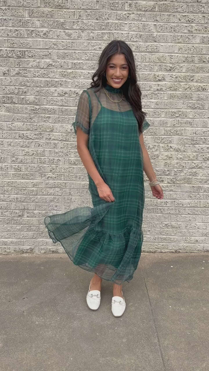 Model standing outside wearing the Glenhaven Plaid Ruffle Maxi Dress in emerald green and navy featuring a high neckline, sheer short sleeves, relaxed silhouette, and flowy ruffled hem. Southern House Clothing
