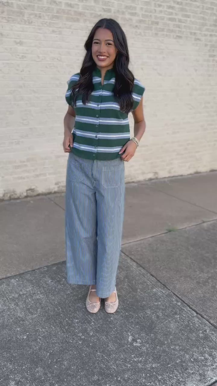 A woman wearing a green and white striped short-sleeve knit top, light blue wide-leg denim jeans, a chunky orange beaded necklace, and beige flats, standing outdoors against a light brick wall.