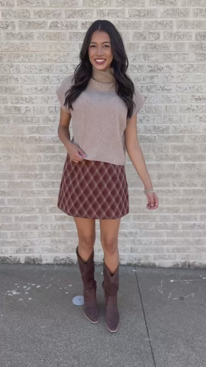 A woman stands on a sidewalk wearing a beige short-sleeve mock neck top, a brown and red plaid mini skirt, and dark brown cowboy boots, posing in front of a white brick wall.