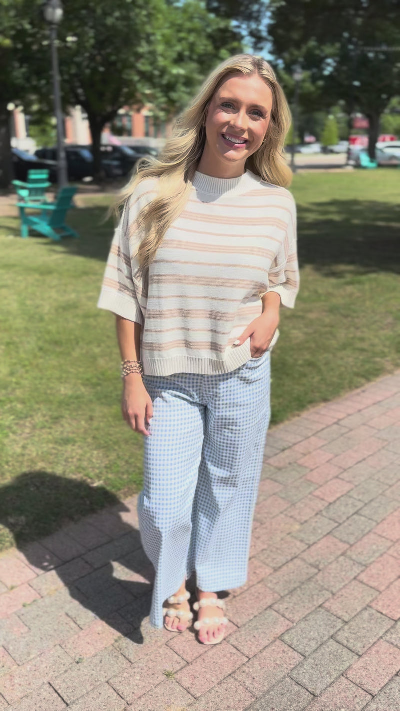 Blonde model wearing a taupe knit top and blue gingham pants, standing outside.
