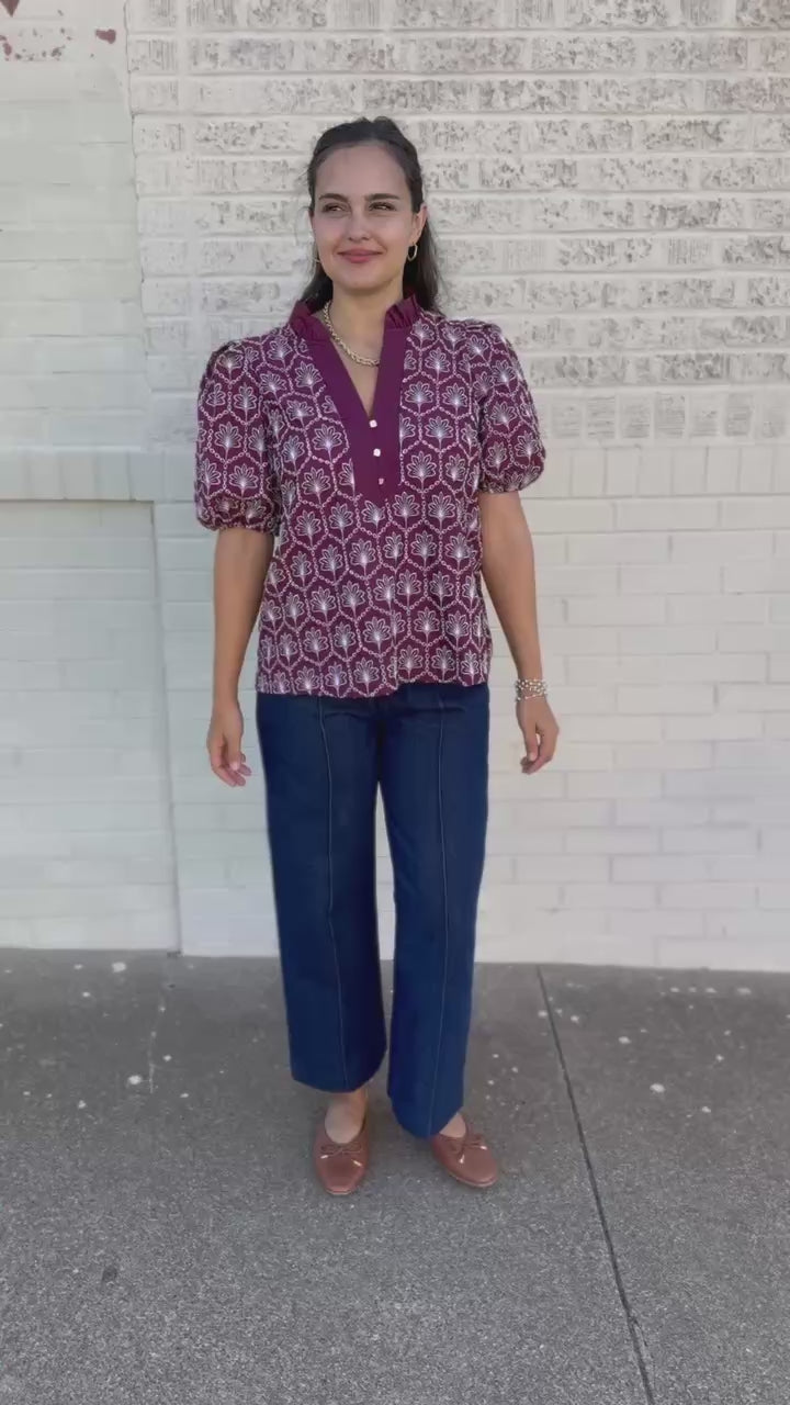 A woman walking outdoors on a sidewalk wearing a short-sleeve button-front blouse with a purple, pink, and white geometric pattern, paired with dark wide-leg pants and brown loafers.
