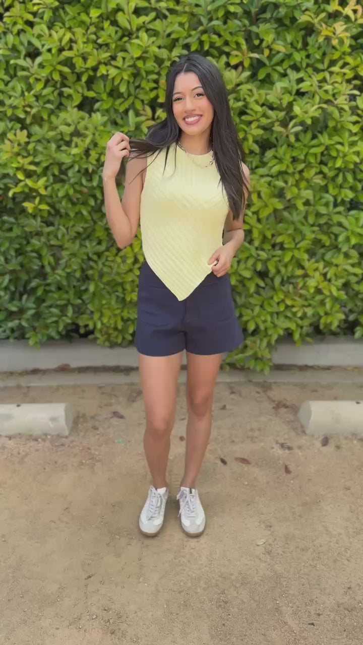 Woman wearing a yellow sleeveless top and blue shorts standing outdoors.