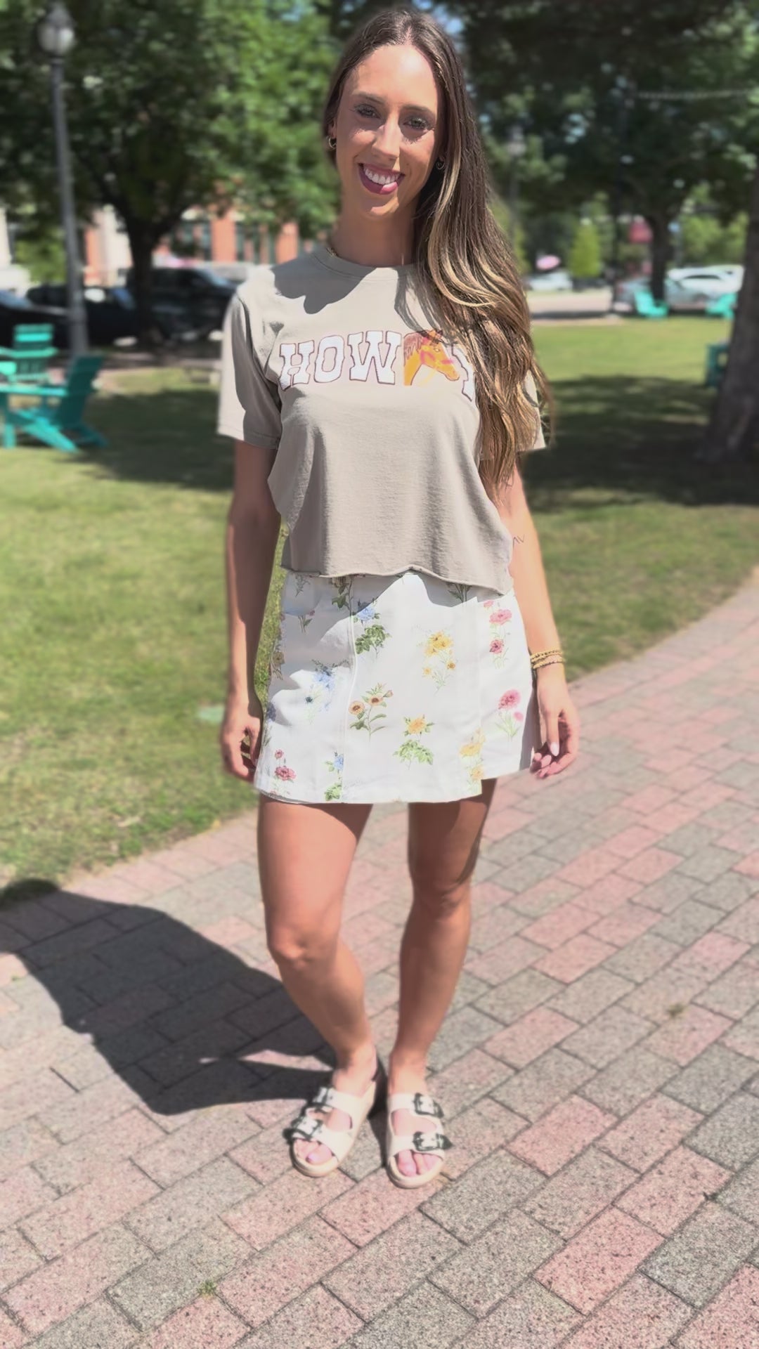 A woman wearing a light tan graphic t-shirt with 'HOWDY' and a peach slice graphic, paired with a white floral mini skirt.