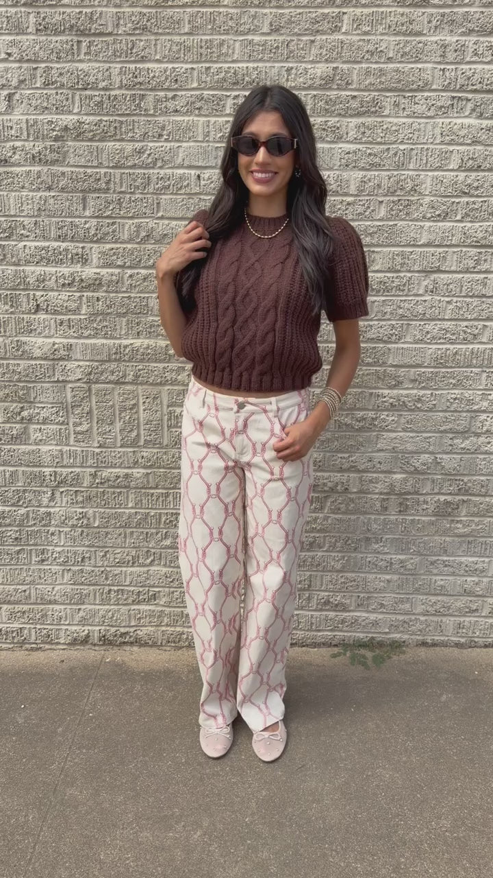 Brunette model wearing the Huxley Cable Knit Sweater Top in Dark Chocolate featuring a textured cable knit pattern, ribbed crew neckline, and short puff sleeves, paired with the Equestrian Charm Horseshoe Print Pants in Ivory and Pink styled, featuring an allover pink rope and horseshoe design. Southern House Clothing