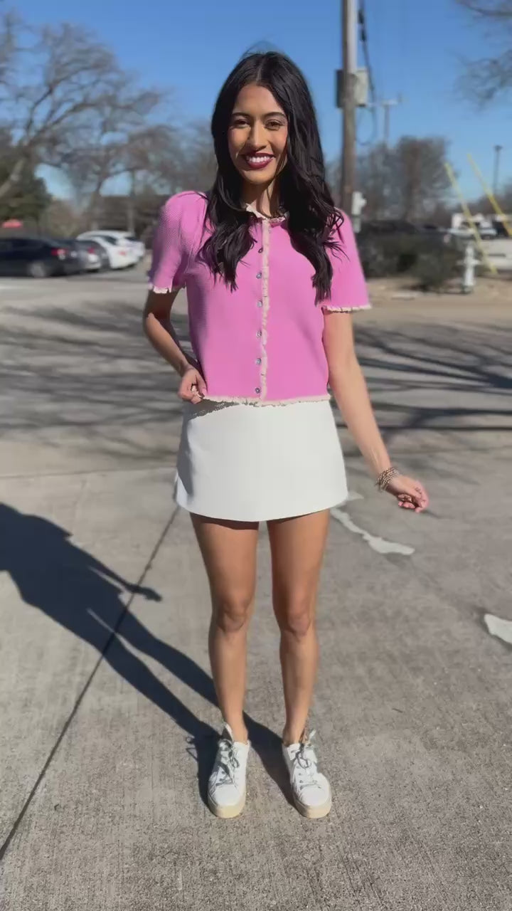 Woman wearing a pink blouse and white skirt standing outside.