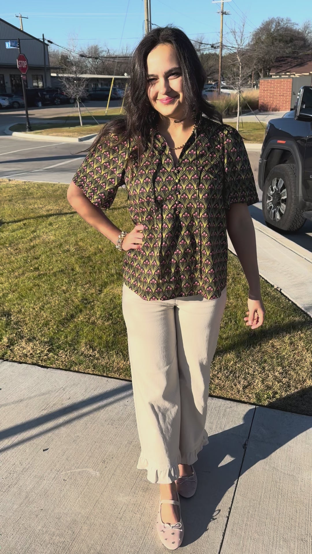 Woman wearing a patterned blouse and beige pants standing outdoors.