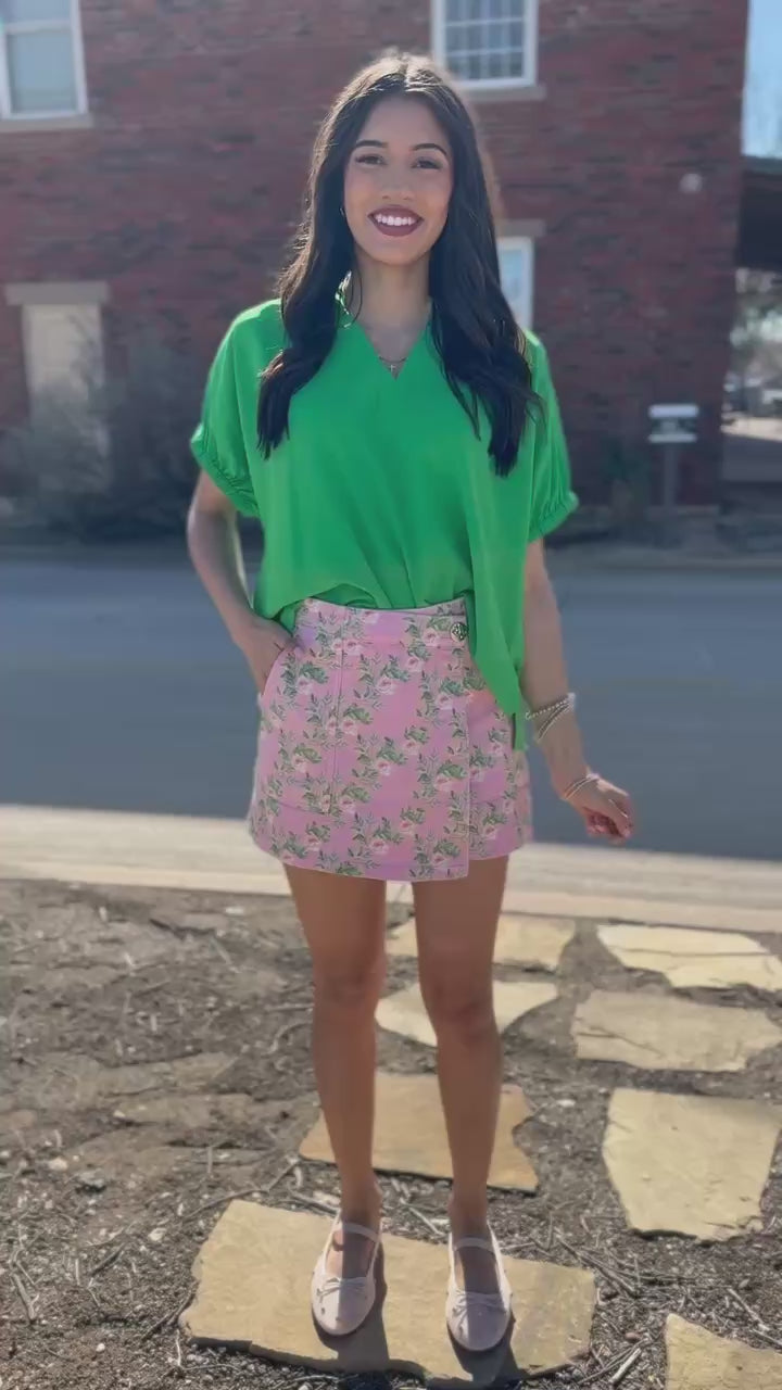 Woman wearing a green blouse and pink floral skirt standing next to a building.