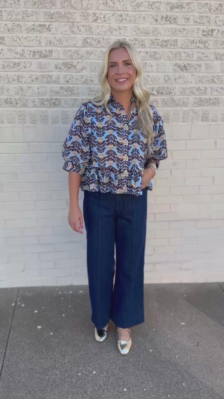 A smiling blonde woman wearing a brown, beige, and white patterned blouse with dark blue wide-leg jeans, standing outdoors on a sidewalk next to a building.